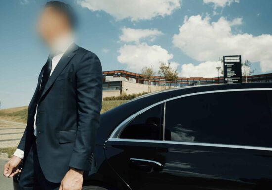 Bodyguard inhuren | Infinite Risks | De afbeelding toont een persoon in een donker pak die naast een zwarte luxe auto staat, met op de achtergrond een modern gebouw en een deels bewolkte lucht. Het gezicht van de persoon is vervaagd, wat privacy of anonimiteit suggereert. De scène speelt zich buiten af op een zonnige dag met een geplaveide ruimte en een grashelling zichtbaar op de achtergrond.