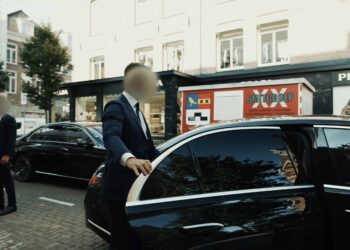 Close protection | Infinite Risks | Two security personnel in formal suits stand next to a black luxury car parked on an urban street, with one holding the car door open.