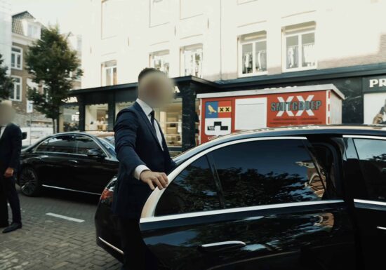 Close protection | Infinite Risks | Two security personnel in formal suits stand next to a black luxury car parked on an urban street, with one holding the car door open.