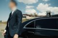 Executive Protection | Infinite Risks | Businessman in a suit standing next to a black luxury car under a blue sky.